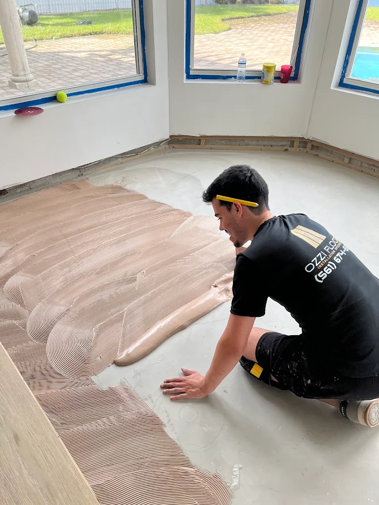 An Ozzifloors installer kneeling on a bare concrete slab, spreading trowel-combed compound ahead of the first row of plank, with the baseboard off and the windows taped